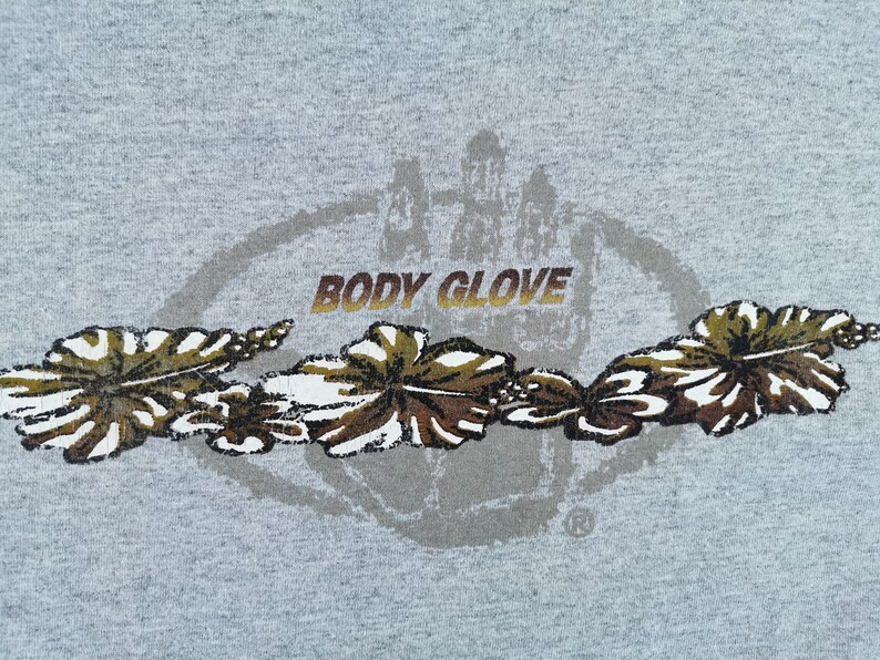 May include: A gray t-shirt featuring the "BODY GLOVE" logo in gold lettering, with a circular design and a handprint graphic. Below the logo are stylized brown and white floral designs.