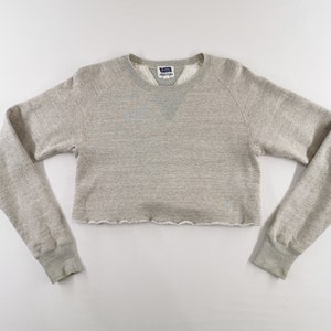 May include: Cropped light grey sweatshirt with long sleeves. The crew neck sweatshirt has a raw hem and a small tag on the neckline. The sleeves have ribbed cuffs. The garment is made of a soft, comfortable material.