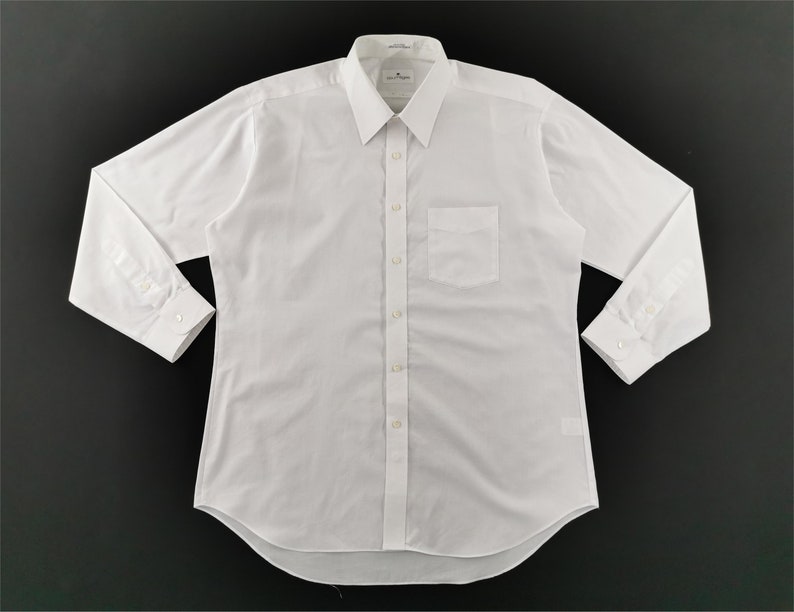 May include: A crisp, white long-sleeve dress shirt with a classic collar and a single chest pocket. The shirt is buttoned up and has buttoned cuffs. The fabric appears to be a smooth, woven material.