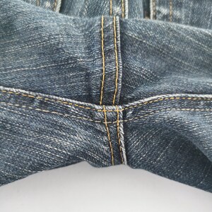 K&ouml;nnte beinhalten: Eine dunkelblaue Jeans aus Denim mit einer markanten Schrittnaht und dicken, kontrastierenden goldgelben Steppn&auml;hten. Der Stoff weist eine subtile, gewaschene Textur und einen getragenen Look auf, was die robuste Verarbeitung des Kleidungsst&uuml;cks unterstreicht.