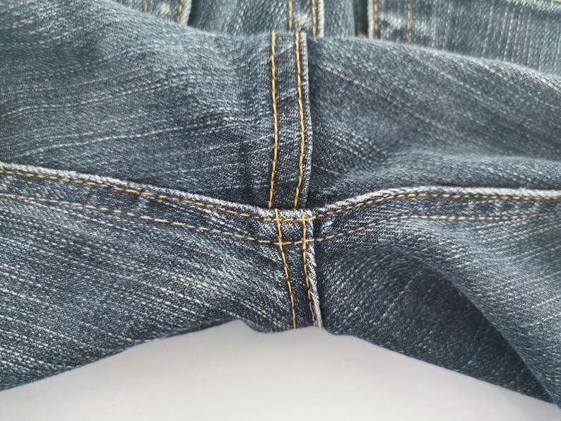 K&ouml;nnte beinhalten: Eine dunkelblaue Jeans aus Denim mit einer markanten Schrittnaht und dicken, kontrastierenden goldgelben Steppn&auml;hten. Der Stoff weist eine subtile, gewaschene Textur und einen getragenen Look auf, was die robuste Verarbeitung des Kleidungsst&uuml;cks unterstreicht.
