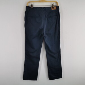 May include: Navy blue pants hanging on a wooden hanger. The pants have a straight leg design and a leather patch on the back. The pants are made of a smooth fabric and appear to be new.