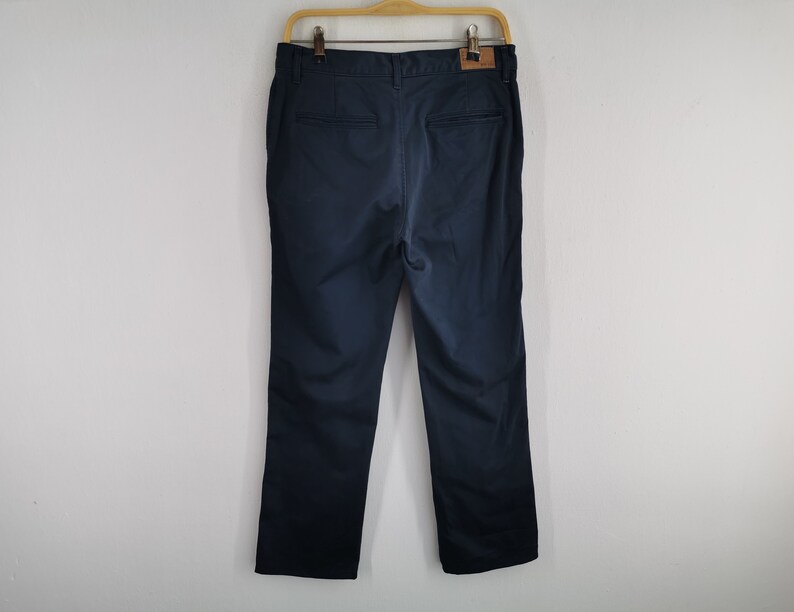 May include: Navy blue pants hanging on a wooden hanger. The pants have a straight leg design and a leather patch on the back. The pants are made of a smooth fabric and appear to be new.