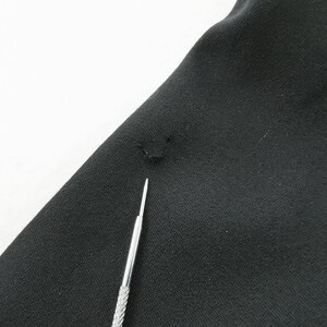 May include: Close-up of black fabric with a small hole and a silver tool. The fabric is slightly wrinkled and appears to be a piece of clothing. The tool is thin and has a textured handle.