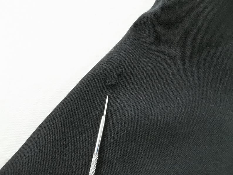 May include: Close-up of black fabric with a small hole and a silver tool. The fabric is slightly wrinkled and appears to be a piece of clothing. The tool is thin and has a textured handle.