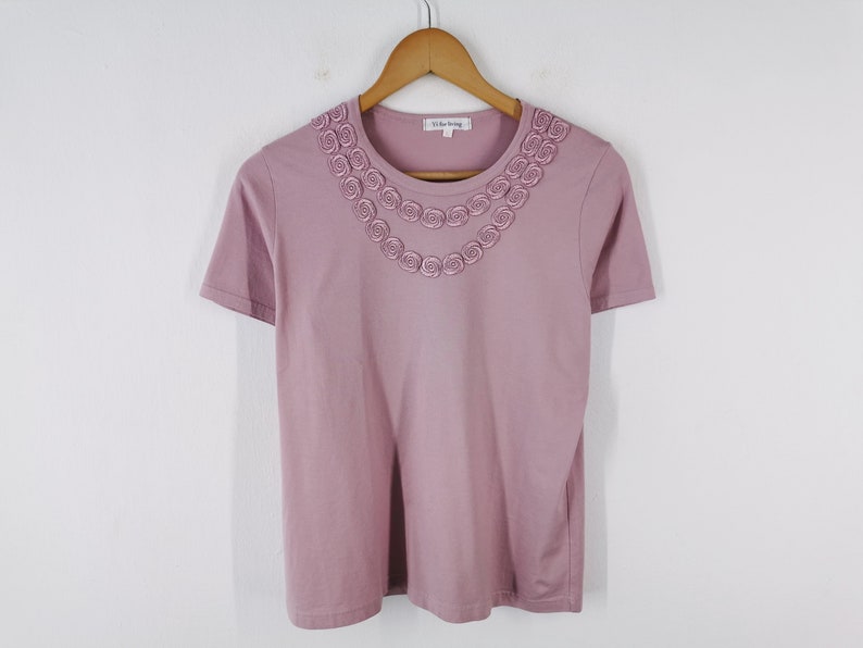 May include: Mauve-colored short-sleeved t-shirt with a decorative floral design around the neckline. The shirt is hanging on a wooden hanger against a white background. The brand name "V. Day Spring" is visible on the label.