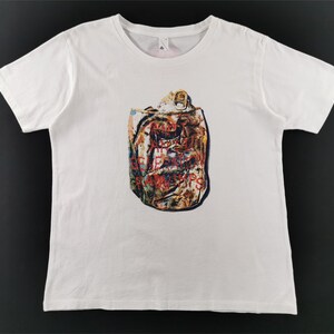 Puede incluir: Una camiseta blanca de manga corta con un estampado gráfico central de una lata de refresco aplastada y multicolor. El diseño muestra una textura metálica desgastada con tonos rojos, azules y dorados, presentando una representación realista de un envase de bebida de aluminio arrugado sobre un fondo de tela blanca lisa.