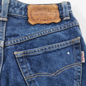 May include: Close-up of blue denim jeans with visible stitching and a brown leather patch. The patch reads "SOMETHING" and "EDWIN". A small tag with the word "SOMETHING" is on the back pocket. The jeans are a classic style.