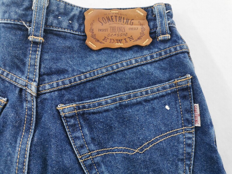 May include: Close-up of blue denim jeans with visible stitching and a brown leather patch. The patch reads "SOMETHING" and "EDWIN". A small tag with the word "SOMETHING" is on the back pocket. The jeans are a classic style.