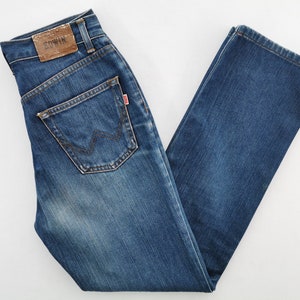 May include: Blue denim jeans with a classic five-pocket design. Features a brown leather patch with the brand name and a red tag on the back pocket. The jeans have a straight leg cut and a faded wash.