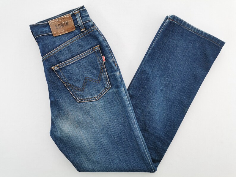 May include: Blue denim jeans with a classic five-pocket design. Features a brown leather patch with the brand name and a red tag on the back pocket. The jeans have a straight leg cut and a faded wash.