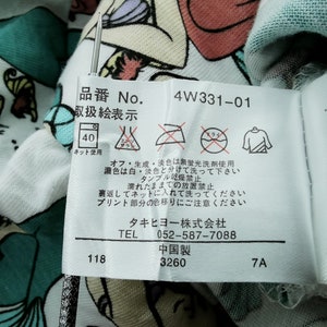 May include: Close-up of a white fabric with a colorful cartoon design. A tag with Japanese text and care symbols is attached. The tag includes the number "4W331-01" and washing instructions.