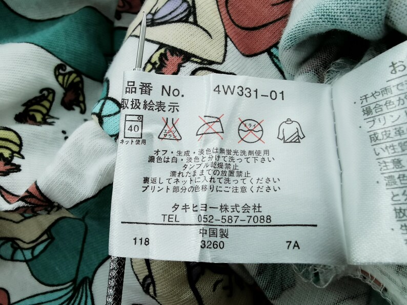 May include: Close-up of a white fabric with a colorful cartoon design. A tag with Japanese text and care symbols is attached. The tag includes the number "4W331-01" and washing instructions.