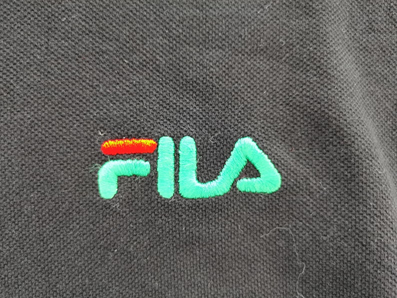 May include: Close-up of a black polo shirt with the embroidered FILA logo. The logo features the word "FILA" in a vibrant green, with the "F" and "A" in a bright green color and the "I" and the horizontal line of the "L" in red and orange.