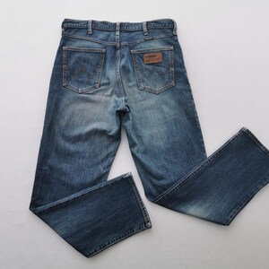 May include: Blue denim jeans with a classic five-pocket design. The jeans feature a faded wash and a brown leather patch with the brand name on the back. The jeans are laid flat on a white surface.