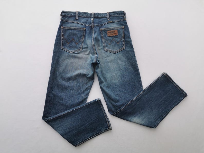 May include: Blue denim jeans with a classic five-pocket design. The jeans feature a faded wash and a brown leather patch with the brand name on the back. The jeans are laid flat on a white surface.