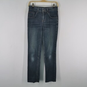 K&ouml;nnte beinhalten: Eine dunkelblaue Jeans aus Denim mit klassischem F&uuml;nf-Taschen-Design, Knopf- und Rei&szlig;verschluss, G&uuml;rtelschlaufen und dezenter Waschung an den Oberschenkeln, die an einem Holzkleiderb&uuml;gel vor einem hellen Hintergrund h&auml;ngt.