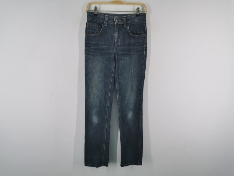 K&ouml;nnte beinhalten: Eine dunkelblaue Jeans aus Denim mit klassischem F&uuml;nf-Taschen-Design, Knopf- und Rei&szlig;verschluss, G&uuml;rtelschlaufen und dezenter Waschung an den Oberschenkeln, die an einem Holzkleiderb&uuml;gel vor einem hellen Hintergrund h&auml;ngt.