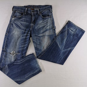 K&ouml;nnte beinhalten: Blaue Jeans aus Denim mit Used-Details, einschlie&szlig;lich eines zerrissenen Knies und verblichenen Patches. Die Jeans haben ein klassisches F&uuml;nf-Taschen-Design und eine gerade Beinpassform. Der Denim ist eine mittelblaue Waschung mit dezenten Whisker-Effekten.