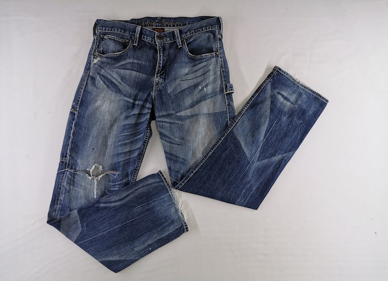 K&ouml;nnte beinhalten: Blaue Jeans aus Denim mit Used-Details, einschlie&szlig;lich eines zerrissenen Knies und verblichenen Patches. Die Jeans haben ein klassisches F&uuml;nf-Taschen-Design und eine gerade Beinpassform. Der Denim ist eine mittelblaue Waschung mit dezenten Whisker-Effekten.