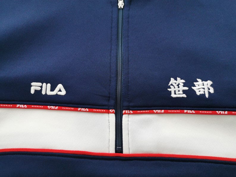 May include: Navy blue athletic jacket with a white stripe and red trim. The jacket features the FILA logo in white on the left chest and Japanese characters on the right chest. A full-length zipper runs down the center.