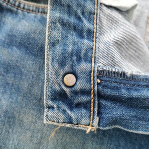Puede incluir: Primer plano de unos vaqueros azules con un bot&oacute;n met&aacute;lico. Los vaqueros muestran costuras detalladas y un aspecto desgastado. El bot&oacute;n es de color plateado y tiene un dise&ntilde;o circular.
