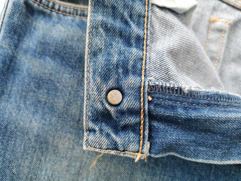 Puede incluir: Primer plano de unos vaqueros azules con un bot&oacute;n met&aacute;lico. Los vaqueros muestran costuras detalladas y un aspecto desgastado. El bot&oacute;n es de color plateado y tiene un dise&ntilde;o circular.