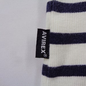 May include: A ribbed fabric garment featuring horizontal navy blue and white stripes. A small black rectangular tag is sewn onto the side, displaying the white text AVIREX with a registered trademark symbol positioned vertically on the fabric.