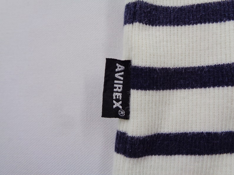 May include: A ribbed fabric garment featuring horizontal navy blue and white stripes. A small black rectangular tag is sewn onto the side, displaying the white text AVIREX with a registered trademark symbol positioned vertically on the fabric.