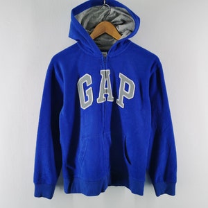 Könnte beinhalten: Ein leuchtend blauer Kapuzenpullover mit Reißverschluss und dem Wort GAP in großen grauen Buchstaben auf der Brust. Das Kleidungsstück hat eine Kapuze mit grauem Innenfutter, lange Ärmel mit gerippten Bündchen und zwei Vordertaschen aus Textilstoff.