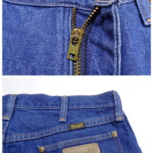 May include: Close-up of blue denim jeans. The jeans feature a brass zipper, a button closure, and the Wrangler logo on a brown leather patch. The stitching is in a contrasting yellow-brown color.