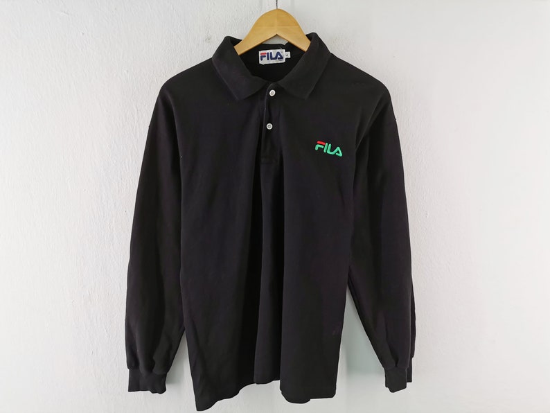 May include: Black long-sleeve polo shirt with a collar and three buttons. The shirt features the FILA logo in green and red on the left chest and the FILA logo on the collar. The shirt is hanging on a wooden hanger.