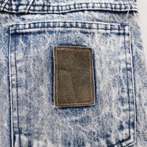 May include: Close-up of a section of acid wash denim jeans. The jeans have a light blue and white pattern with a brown rectangular patch on the back pocket. The stitching is visible.