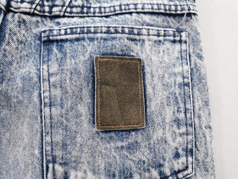 May include: Close-up of a section of acid wash denim jeans. The jeans have a light blue and white pattern with a brown rectangular patch on the back pocket. The stitching is visible.