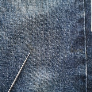 K&ouml;nnte beinhalten: Nahaufnahme von blauem Denim-Stoff mit einem silbernen Metallwerkzeug. Der Denim hat ein strukturiertes Aussehen mit subtilen Farbvariationen. Der Stoff ist ein klassisches Blue-Jeans-Material.