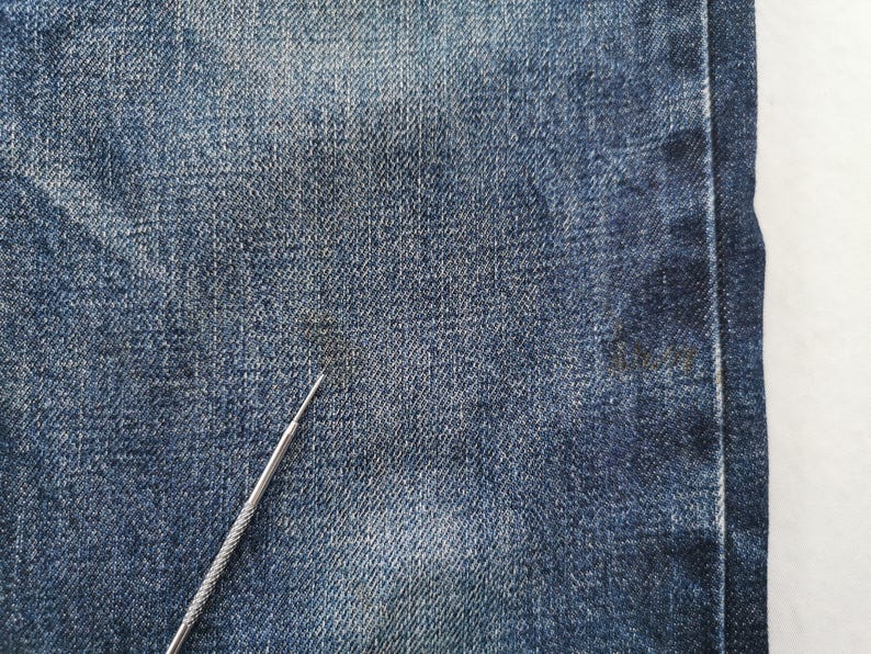 K&ouml;nnte beinhalten: Nahaufnahme von blauem Denim-Stoff mit einem silbernen Metallwerkzeug. Der Denim hat ein strukturiertes Aussehen mit subtilen Farbvariationen. Der Stoff ist ein klassisches Blue-Jeans-Material.