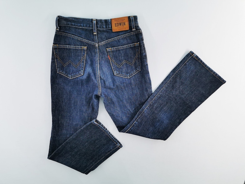May include: Dark blue denim jeans with a classic five-pocket design. Features a brown leather patch with the brand name "EDWIN" on the back waistband. The back pockets have a distinctive W-shaped stitch detail. The jeans are laid flat.