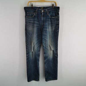 May include: Blue denim jeans with a faded wash and distressed details, hanging on a wooden hanger. The jeans have a classic five-pocket design and a button and zipper closure. The jeans are a size 36.
