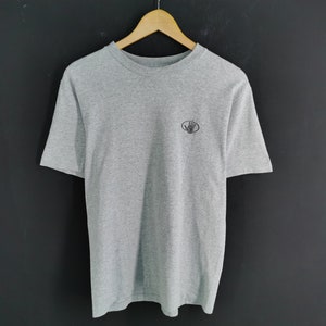 May include: A heather gray t-shirt with short sleeves, hanging on a wooden hanger. The shirt has a small, dark logo on the upper left chest. The background is a dark gray.