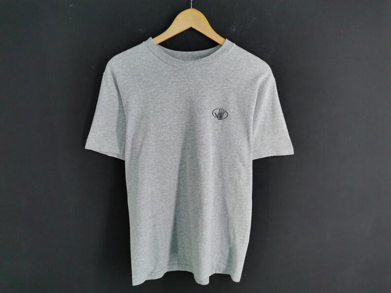 May include: A heather gray t-shirt with short sleeves, hanging on a wooden hanger. The shirt has a small, dark logo on the upper left chest. The background is a dark gray.