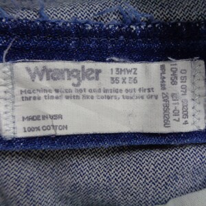May include: Close-up of a blue denim fabric with a white Wrangler tag. The tag displays the size 35 x 36, along with washing instructions and the text "MADE IN USA" and "100% COTTON".