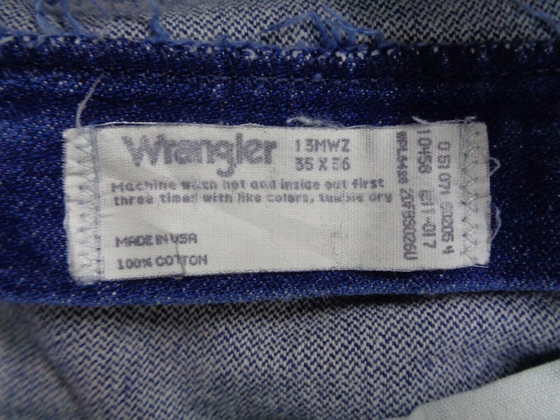 May include: Close-up of a blue denim fabric with a white Wrangler tag. The tag displays the size 35 x 36, along with washing instructions and the text "MADE IN USA" and "100% COTTON".