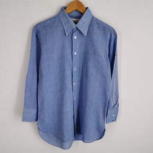 Può includere: Una camicia azzurra con bottoni, maniche lunghe, colletto a punta e taschino sul petto. La camicia è appesa a una gruccia di legno su sfondo bianco. La camicia ha un design classico.