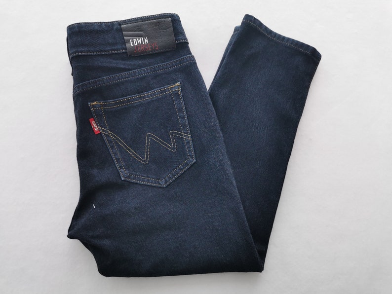 K&ouml;nnte beinhalten: Dunkelblaue Jeans aus Denim mit klassischem F&uuml;nf-Taschen-Design. Mit einem Lederaufn&auml;her mit dem Text "EDWIN" und "JERSEYS" sowie einem roten Logo-Etikett. Die Ges&auml;&szlig;tasche hat ein einzigartiges Nahtdesign.