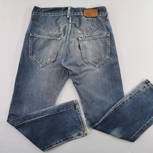 May include: Faded blue denim jeans with a distressed appearance. Features include a brown leather patch, red tag, and distinctive pocket design. The jeans are laid flat, displaying the back pockets and overall style.