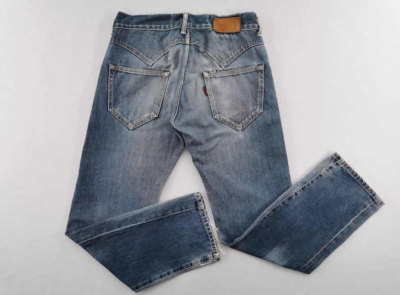 May include: Faded blue denim jeans with a distressed appearance. Features include a brown leather patch, red tag, and distinctive pocket design. The jeans are laid flat, displaying the back pockets and overall style.
