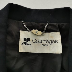 May include: Close-up of a black jacket collar with a label that reads "Courr&egrave;ges paris." The label is cream-colored with black text and a logo. A silver chain is attached to the label.