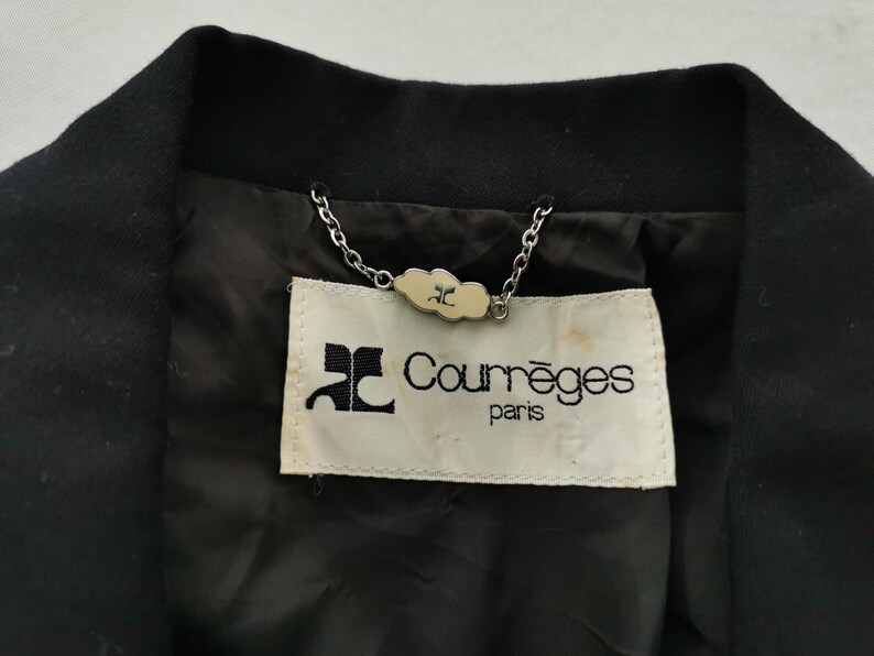 May include: Close-up of a black jacket collar with a label that reads "Courr&egrave;ges paris." The label is cream-colored with black text and a logo. A silver chain is attached to the label.
