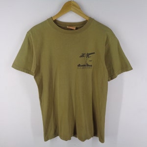 May include: Khaki green t-shirt with a crew neck and short sleeves. The shirt has a small graphic on the upper left chest with the text "C.C. MASTERS" beneath it. The graphic depicts an aeroplane and palm trees.
