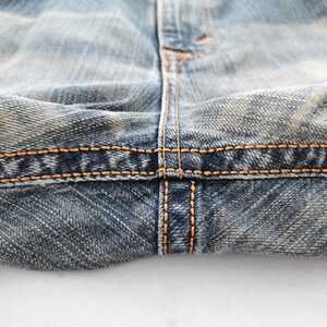 Puede incluir: Primer plano de unos vaqueros de mezclilla azul con costuras en hilo naranja-amarillo. La imagen destaca los detalles de las costuras y la textura de la tela vaquera desgastada.